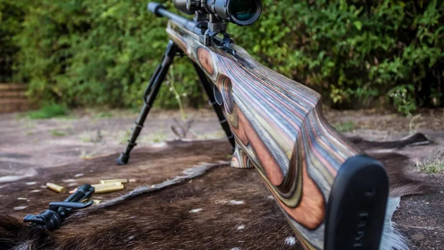 Hunter in camouflage aiming rifle with scope mounted, focusing on distant target in natural woodland setting