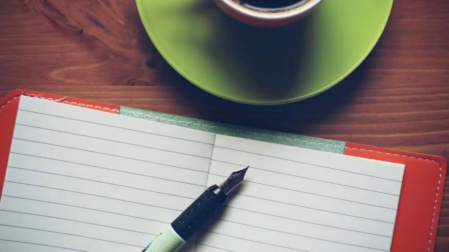 Open notebook lying flat on wooden desk with pen nearby, showing binding and pages