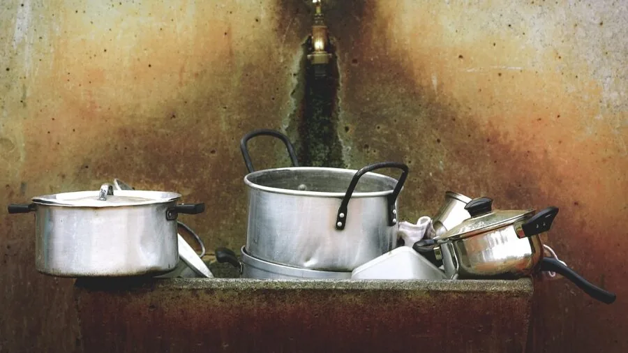 Person cleaning stainless steel pots in a bright kitchen sink with soapy water and a cloth
