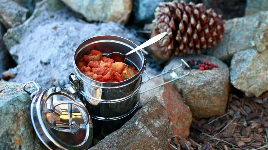 Person holding a metal camping mug by a glowing campfire in natural outdoor setting