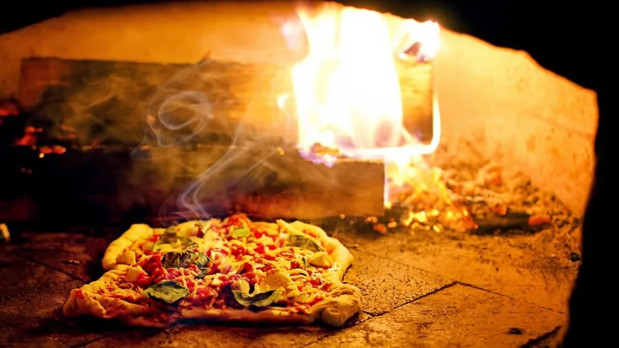 Person preparing fresh pizza dough near traditional outdoor pizza oven with warm lighting