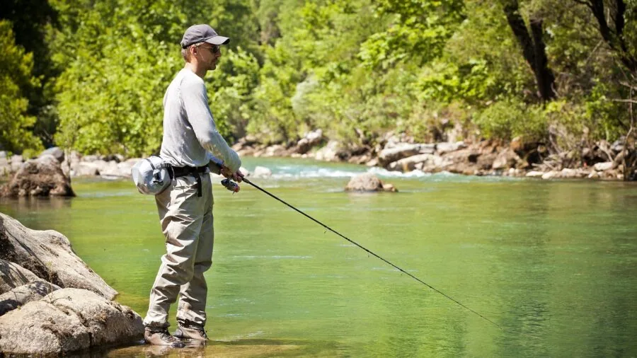 Person wearing fishing vest standing by riverbank preparing fishing gear in outdoor setting