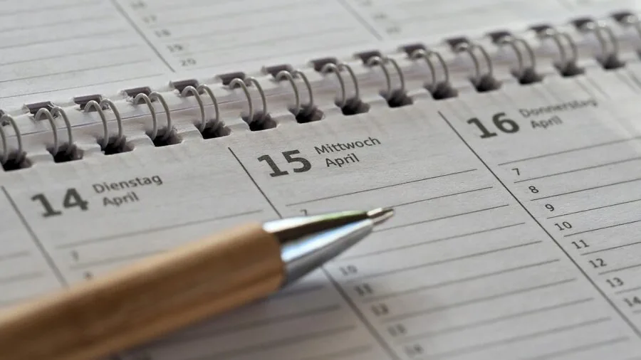 Person writing in planner with calendar and weekly schedule visible on desk