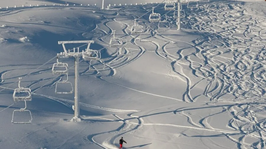 Skier in winter gear adjusting ski boot flex on snowy mountain slope during daytime