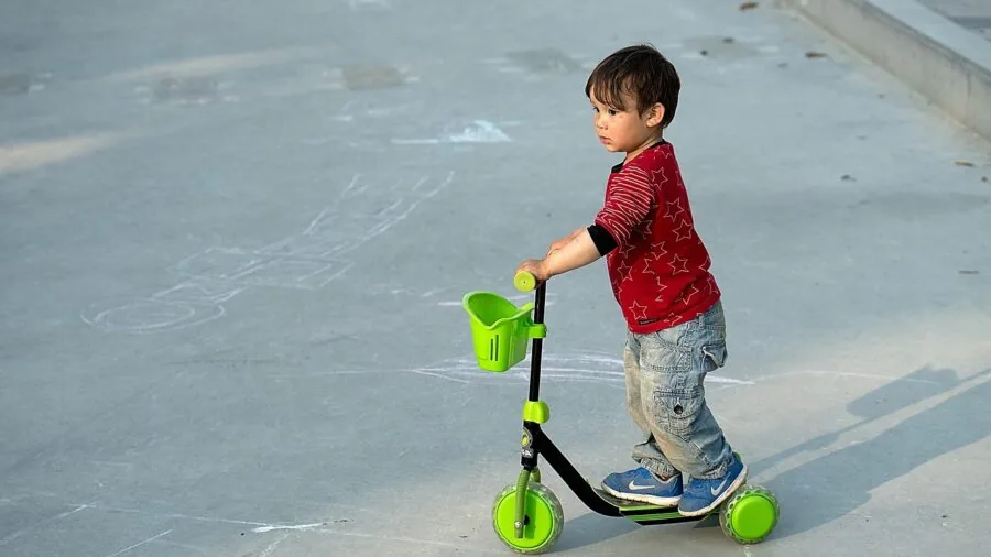 Small child learning to ride a three-wheel scooter on a sunny pathway in a park setting