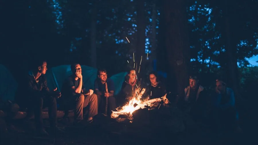 Two friends sitting in camping chairs together by a campfire in a natural outdoor setting