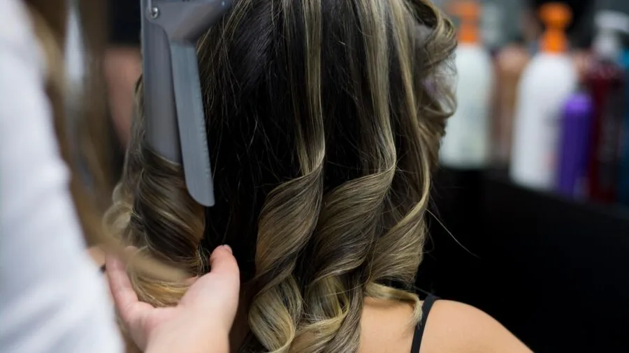 Woman using a curling wand to style her hair in a bright bathroom with natural lighting