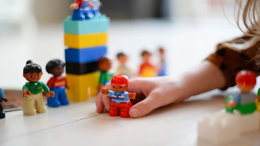 Young child engaged in imaginative doll play with various accessories and clothing items on wooden floor