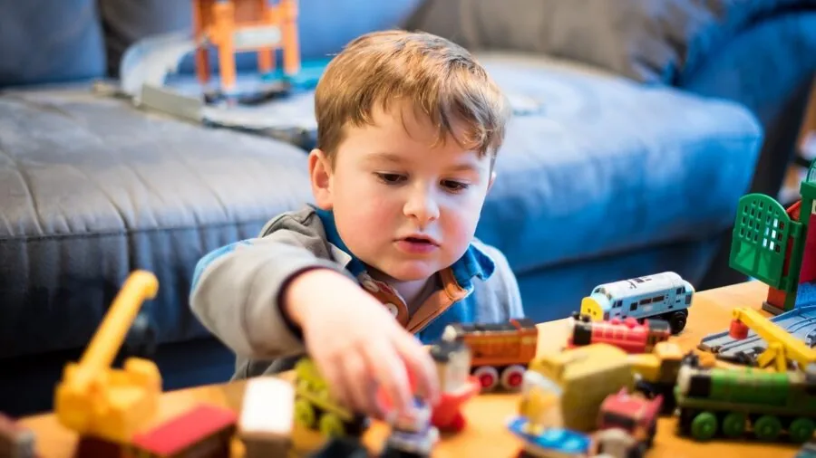 Young child engaged with colorful toy train set on wooden floor, demonstrating imaginative play and investment in toys.