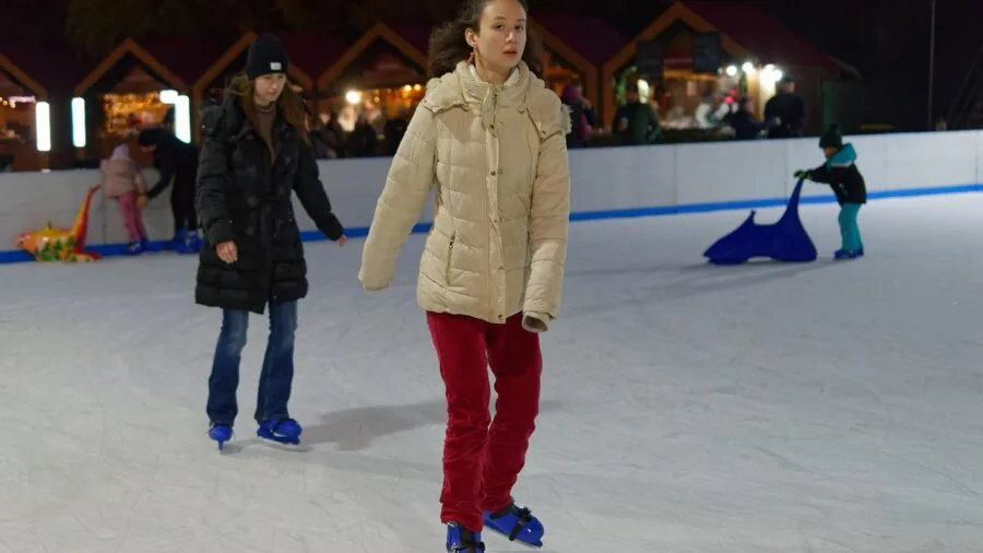 Young child in winter clothes learning to ice skate on frozen pond with adult supervision
